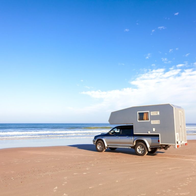 absetzkabine-pickup-camping-am-strand. Pickup mit Wohnaufbau am Strand, sanftes Meer im Hintergrund unter blauem Himmel.